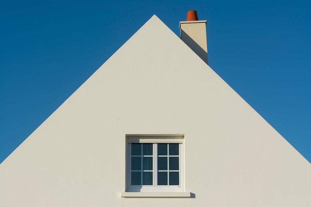 Simple triangular house facade with window and chimney against a clear blue sky.